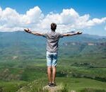 A man stands triumphantly with arms open, overlooking a scenic mountain range.