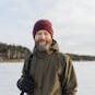 A man smiles while ice fishing on a frozen lake, dressed warmly for winter.